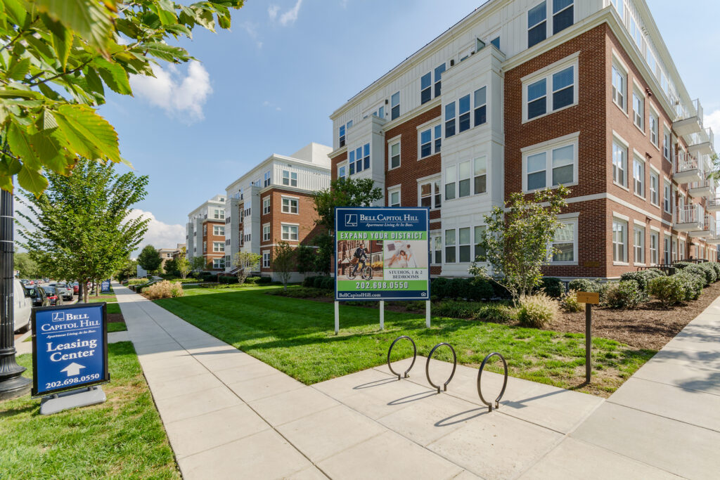 A modern apartment complex with red brick and white trim, surrounded by green lawns and trees. Signs for Leasing Center and Bell Capitol Hill are visible near the sidewalk on a sunny day.
