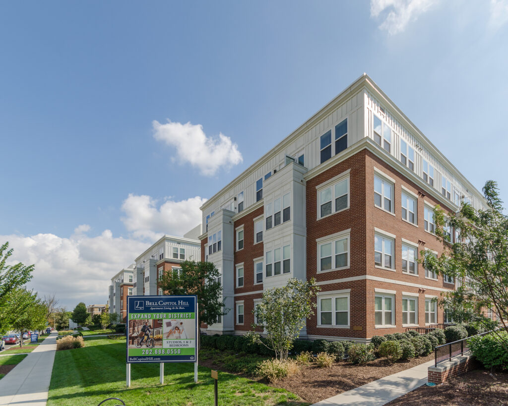 A modern, multi-story apartment building with red brick and white siding, surrounded by landscaping. A sign in the foreground advertises leasing for Bell Cherry Hill. The sky is mostly clear with a few clouds.