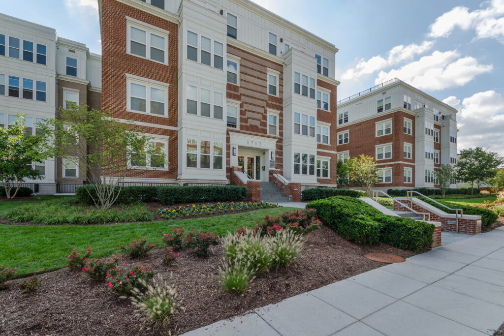 A modern, multi-story apartment building with red brick and white accents, landscaped grounds, shrubs, flowers, and a sidewalk under a partly cloudy sky.