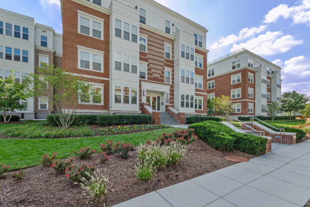 A modern apartment building with red brick and white siding, landscaped with bushes, flowers, and green grass, under a partly cloudy sky. Steps and walkways lead to the entrance.